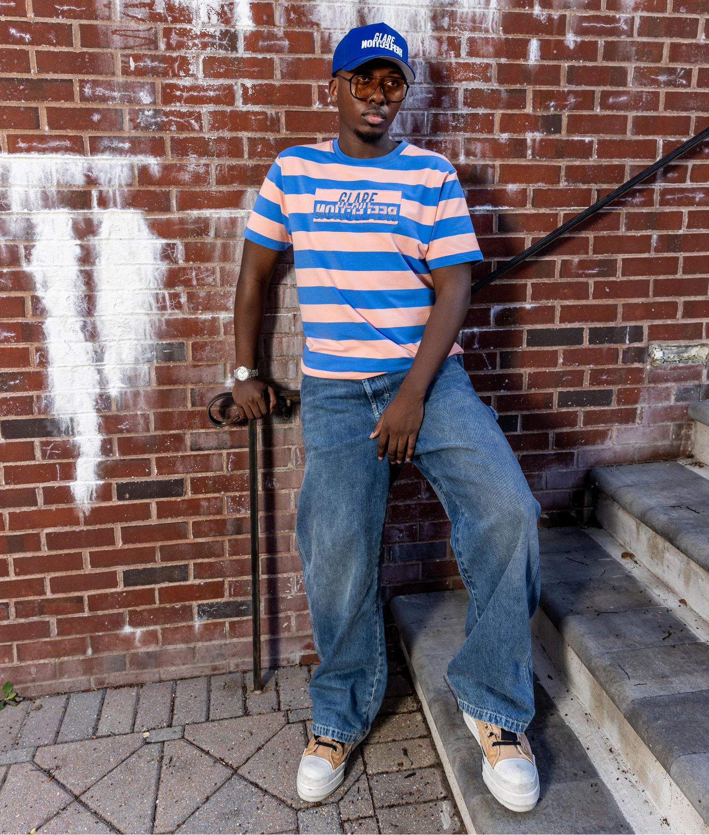 Person wearing a striped shirt and jeans sitting against a brick wall.